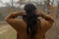 Young woman with long dark hair wearing brown knit sweater standing outdoors in autumn setting, view from behind as she touches her hair, creating atmospheric mood.