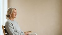 Mature woman with gray hair in casual sweater sitting alone by window, looking thoughtful and contemplative in bright natural light.