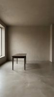 Minimalist interior space featuring a single wooden table in an empty room with concrete flooring, neutral walls, and natural light from windows, suitable for architectural and design projects.