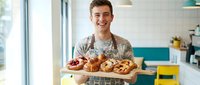 Young Caucasian male baker wearing apron holding wooden board with fresh baked croissants and pastries in modern kitchen, suitable for bakery, food business, and culinary content.