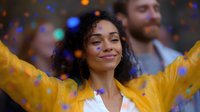 Young African American woman in yellow jacket celebrating with arms raised, expressing joy and freedom amid colorful bokeh lights and confetti. Perfect for lifestyle content.