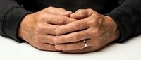 Senior businessman hands clasped together on white desk surface showing wedding ring and professional attire in office meeting setting.