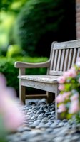 Weathered wooden bench in tranquil garden setting with smooth pebbles and soft pink flowers, creating peaceful outdoor relaxation space.