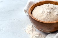 Fresh white wheat flour in wooden bowl on light background, scattered powder on rustic table with linen napkin, baking and cooking ingredients.