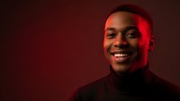 Young African American man smiling confidently in black turtleneck with dramatic red lighting against dark background for professional headshot.