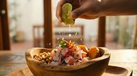 Young adult male hand squeezing fresh lime juice over ceviche bowl with seafood, potatoes and vegetables in wooden serving dish, bright kitchen setting.