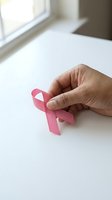 Hand holding pink breast cancer awareness ribbon on white surface near window for healthcare campaign and support initiatives.
