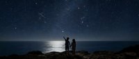 Romantic couple silhouettes stargazing under starry night sky by moonlit ocean. Two people enjoying celestial view on rocky coastline during evening.