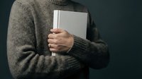 Person in cozy knit sweater holding blank notebook against dark background. Minimalist lifestyle concept for journaling, writing, education projects.