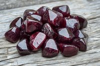 Polished dark red garnet crystals scattered on rustic wooden surface, showing natural mineral formation with deep burgundy color and glossy facets in macro view.