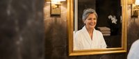 Mature woman with gray hair smiling in luxury bathroom mirror wearing white bathrobe enjoying spa wellness routine for skincare and self-care relaxation.