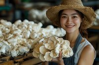 Young Asian woman farmer in straw hat holding fresh white mushrooms at organic farm, displaying harvest with genuine smile, wearing casual attire.