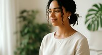 Young woman with dark curly hair and medium skin tone wearing white casual t-shirt, smiling indoors near green plants, suitable for lifestyle, wellness, and portrait stock use.