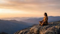 Woman meditating on mountain peak during golden hour sunrise with layered mountain ranges in background creating peaceful mindfulness scene for wellness content.