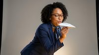 African American businesswoman in navy suit and glasses throwing paper airplane in office setting for corporate leadership and innovation concepts.