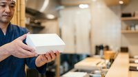 Middle-aged male with light skin tone, wearing dark blue chef's coat, holding white takeout box in modern restaurant kitchen with blurred background and warm lighting.