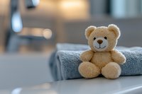 Soft plush teddy bear sitting on folded gray towel with blurred bathroom background, creating cozy and gentle atmosphere for childcare and comfort themes.