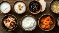 Variety of healthy breakfast toppings in ceramic bowls on wooden table, including yogurt, granola, seeds, nuts and dried fruits. Top view food styling.