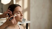 Professional makeup artist applying foundation to young woman's face with brush in bright studio setting for beauty treatment session.
