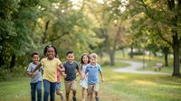 Group of five diverse children running together on tree-lined park path during sunny day, enjoying outdoor recreation and friendship activities.