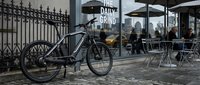 Modern hybrid bicycle parked on urban street near coffee shop with outdoor seating area and ornate metal fence, suitable for lifestyle and transportation concepts.