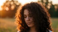 Young mixed-race woman with curly hair in golden hour sunlight, peaceful expression, warm outdoor portrait for lifestyle and wellness content.