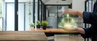 Young professional in dark business attire holding glowing holographic house icon above wooden desk in modern office with plants and windows, representing smart home technology and real estate investment.