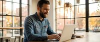 Smiling bearded businessman working on laptop in modern office with large windows during golden hour light for remote work and productivity concepts.