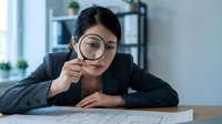 Young Asian businesswoman examining documents with magnifying glass at office desk, focused on detailed analysis and investigation work.