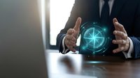 Professional businessman in dark suit holding glowing digital compass hologram between hands at office desk for business direction and strategy concepts.