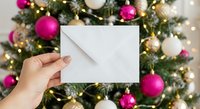 Hand holding white envelope against decorated Christmas tree with pink, gold and white ornaments and warm lights for holiday greeting cards and festive correspondence.