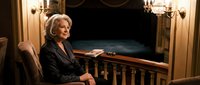 Elegant senior Caucasian businesswoman in formal black suit sitting confidently at wooden desk in luxurious office interior with chandelier lighting.