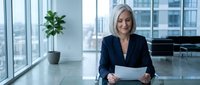Mature woman with gray hair and light complexion, wearing professional dark blazer, holding tablet in modern high-rise office with city skyline view, business technology concept.
