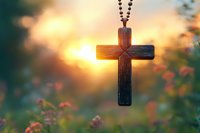 Wooden cross pendant on beaded chain silhouetted against golden sunset with soft-focus wildflowers in foreground, creating spiritual atmosphere.