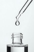 Clear liquid drop suspended from glass dropper above transparent bottle on white background, minimalist black and white composition for cosmetic and pharmaceutical concepts.