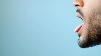 Male profile showing tongue sticking out against light blue background, expressing playful or teasing gesture with stubble beard visible in close-up view.