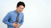 Asian businessman in blue shirt experiencing severe stomach pain holding abdomen with distressed facial expression on white background.