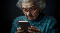 Senior Caucasian woman with gray hair and glasses using smartphone in teal sweater against dark background for technology and aging concepts.