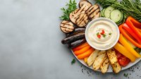 Creamy hummus dip surrounded by colorful grilled vegetables and bread on gray background, perfect for healthy appetizer platter.