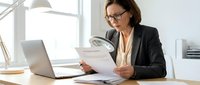 Professional businesswoman in glasses reviewing documents at modern office desk with laptop and desk lamp for corporate presentations.