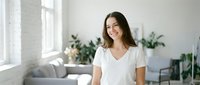 Young Caucasian woman in white t-shirt smiling at camera in bright modern living room with plants and natural light streaming through windows.