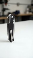 Black folding pocket knife with steel blade standing upright on white surface in modern kitchen setting for outdoor camping and utility use.