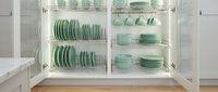 Organized kitchen cabinet with mint green ceramic dishes, plates, bowls, and cups neatly arranged on chrome wire racks with soft lighting.