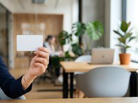Businessman holding blank white business card in modern office workspace with colleagues and green plants in background, suitable for corporate branding and professional networking concepts.