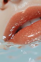 Close-up of glossy coral lips with water droplets and reflections against turquoise background, showing moisturized skin texture and makeup artistry details.