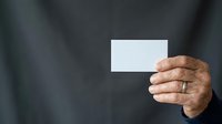 Mature man with medium skin tone holding blank white business card in hand against dark blurred background, suitable for corporate branding and professional networking materials.