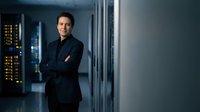 Confident businessman in dark suit standing with crossed arms in modern data center server room with glowing equipment and professional lighting atmosphere.