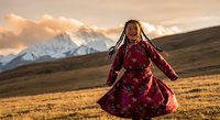 Young Tibetan woman in traditional red dress dancing joyfully in mountain meadow at golden hour sunset with snow-capped peaks in background.