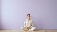 Woman in casual beige clothing sitting cross-legged in meditation pose against purple wall background, promoting wellness, mindfulness and relaxation.