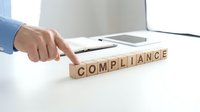 Caucasian male hand in blue business shirt pointing at wooden blocks spelling compliance on white desk with laptop, representing regulatory adherence and business standards.